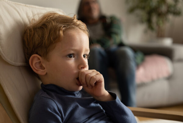 A little boy sits at home in a chair and sucks his finger, in the background a man watches the boy